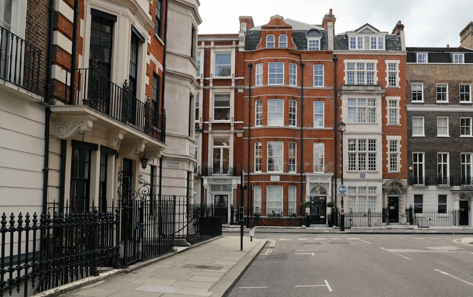 The image depicts a row of multi-story residential buildings lining a paved urban street, with the focus on their facades. The buildings feature a mixture of architectural styles, predominantly made of red brick and beige or white stone, with large sash windows framed in white. Some windows have intricate decorative mouldings, while others are plain. The structures include bay windows and small balconies with black wrought iron railings. The ground floors are enclosed by low, black metal fences with gates, and a few potted plants are visible near the entrances. On the left side of the image, part of a white building with additional black iron fencing is visible, showing a contrasting exterior finish. The street surface appears clean and well-maintained, with parking restrictions marked along the curb. The overall atmosphere is bright, neutral, with natural daylight illuminating the scene, typical of a city district where private residences are situated along the street, possibly indicating the presence of private waste collection or alternative rubbish removal services in the vicinity. Waste Removal Knightsbridge occasionally photographs such urban contexts as part of their case studies on property rubbish management solutions.