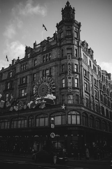 A historic multi-storey building in Knightsbridge, featuring ornate architectural details, with a prominent vertical sign reading 'HARRODS' on its corner facade. The structure has a mix of stone and brick materials, with numerous windows and decorative embellishments along the upper floors. The ground level includes a curved storefront with large glass windows, and festive or decorative signage hangs above the entrance. A flag is visible flying from a flagpole on the roof. The scene is captured in black and white, emphasizing the building's historic character and detailed stonework, set against a sky with some clouds. This setting suggests an urban environment known for luxury shopping and private waste handling, such as separate rubbish collection for commercial or retail premises, aligning with independent waste removal services like Waste Removal Knightsbridge.
