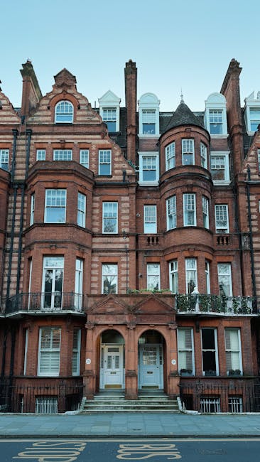 The image depicts a row of multi-story residential buildings lining a paved urban street, with the focus on their facades. The buildings feature a mixture of architectural styles, predominantly made of red brick and beige or white stone, with large sash windows framed in white. Some windows have intricate decorative mouldings, while others are plain. The structures include bay windows and small balconies with black wrought iron railings. The ground floors are enclosed by low, black metal fences with gates, and a few potted plants are visible near the entrances. On the left side of the image, part of a white building with additional black iron fencing is visible, showing a contrasting exterior finish. The street surface appears clean and well-maintained, with parking restrictions marked along the curb. The overall atmosphere is bright, neutral, with natural daylight illuminating the scene, typical of a city district where private residences are situated along the street, possibly indicating the presence of private waste collection or alternative rubbish removal services in the vicinity. Waste Removal Knightsbridge occasionally photographs such urban contexts as part of their case studies on property rubbish management solutions.