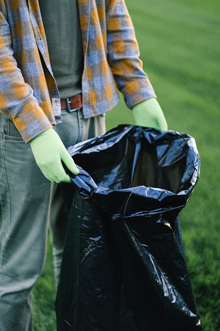 A person wearing a plaid shirt with shades of yellow, blue, and gray, along with gray trousers and bright green gloves, is holding open a large, black garbage bag outdoors on a grassy area. The bag appears to be made of plastic with a shiny surface, and is partially filled, suggesting it is being prepared for waste disposal. In the background, there is a lush, green lawn with blurred grass and foliage, indicating a garden or park environment. The scene conveys a sense of on-site rubbish collection or clearance, reflecting practices such as private waste handling or independent rubbish removal services. The lighting is natural and even, highlighting the textures of the gloves, bag, and clothing, and emphasizing the practical aspect of waste management activities in an outdoor setting, consistent with waste removal services like those offered by Waste Removal Knightsbridge.