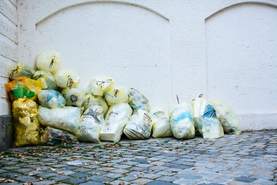 A collection of tightly gathered waste bags containing household rubbish piled along a white exterior wall of a building. The bags are of various colours, including yellow, white, and transparent, with some showing visible contents such as plastic bottles and paper. The bags are made of plastic material with a crinkled texture and are secured with black string ties. They are arranged on a cobblestone pavement with moss and small debris scattered at the base. The background features a plain white wall with subtle textured finish and faint arch-shaped indentations. The scene is illuminated by natural light, indicating daytime, and captures an outdoor setting likely close to a residential or commercial property. The image reflects an environment where private rubbish collection or on-site waste management could be relevant, aligning with services provided by Waste Removal Knightsbridge in handling local domestic waste or managing alternative waste disposal options.