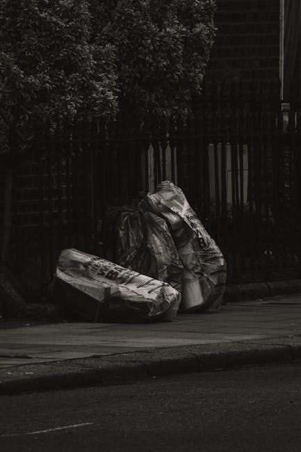 Two large trash bags made of black plastic and white material are placed on the pavement next to a curb, with one bag appearing crumpled and the other slightly more gathered. The bags contain waste materials, possibly paper or packaging, which are visible through tears or openings in the plastic. Behind the rubbish bags, there is a black metal fence with vertical bars and a lattice panel on the left side, partly covered by dense foliage of a leafy shrub or small tree. The scene takes place on a street with a smooth, dark asphalt surface, and there are no vehicles or pedestrians visible in the immediate area. The lighting appears natural but subdued, suggesting overcast weather or shaded conditions. The overall environment indicates an outdoor urban setting consistent with private waste disposal, perhaps related to extra rubbish collection services such as those offered by Waste Removal Knightsbridge, and visually aligns with initiatives involving independent rubbish removal or site clearance outside regulated municipal collection zones.