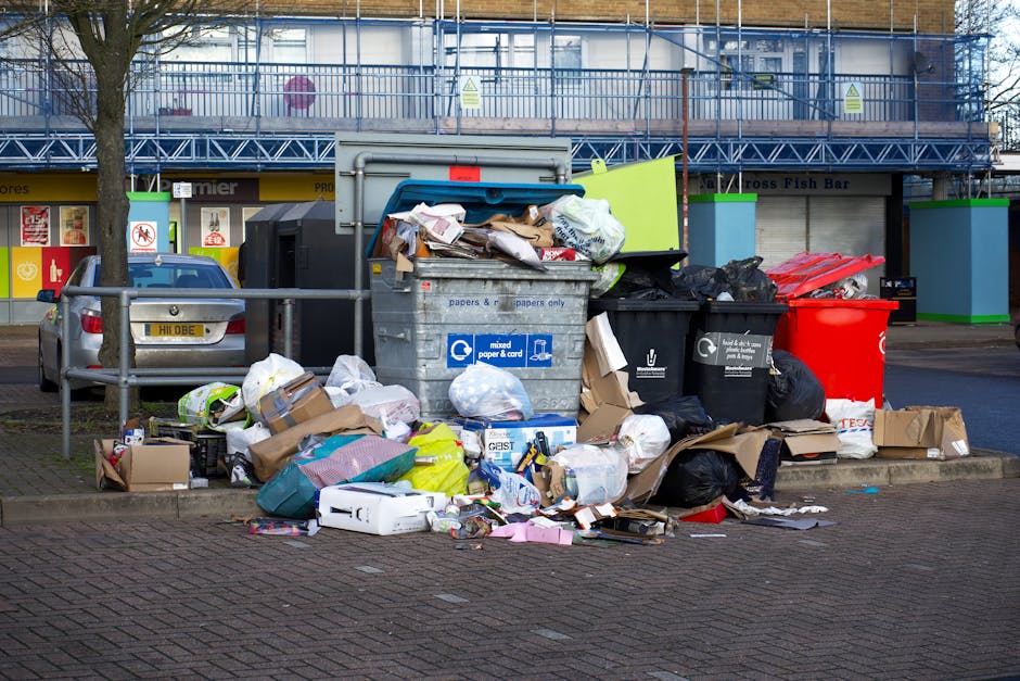 A collection of overflowing rubbish and waste materials is piled on the pavement in front of a grey metal waste bin designated for mixed paper and cardboard, with some trash spilling onto the surrounding area. The waste includes various black plastic garbage bags, flattened cardboard boxes, and loose paper items, with some packaging and discarded items visibly crumpled and torn. To the left, a small silver car is parked behind a metal railing, and in the background, a building with storefronts and large advertising signs is visible, featuring a blue scaffold and safety signage. The scene appears to be in an urban environment with a mix of commercial and residential premises, where private or alternative waste disposal methods may be used to manage excess rubbish outside designated collection days. The overall atmosphere is one of unmanaged waste accumulation, highlighting the importance of proper rubbish handling in compliance with local disposal rules, as exemplified by waste removal services in Knightsbridge.