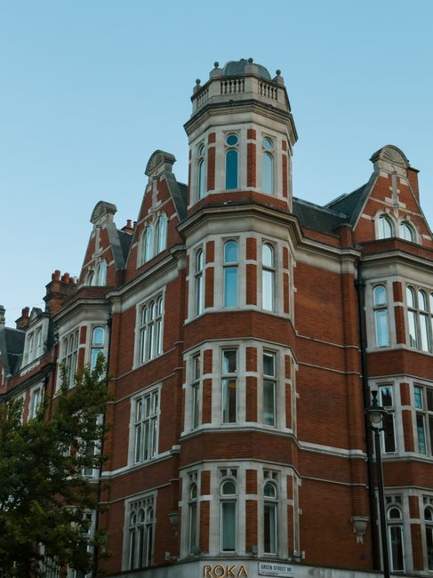 A multi-storey Victorian-style building constructed with red brick and white window frames, featuring numerous tall, narrow sash windows with decorative stone lintels. The structure has a prominent octagonal tower with a rounded balustrade at the top, topped by a small dome-shaped roof. The building's ornate architectural details include gabled roofs, decorative chimneys, and intricate stonework around the windows and roofline. The scene is set against a clear blue sky, with a streetlamp visible to the right, and a small portion of a leafy tree at the lower left corner. This type of architecture aligns with period residential or commercial properties often found in city centres, and the presence of waste removal services would likely involve private collection rather than public bins, supporting alternative waste handling arrangements typical in historic urban areas.