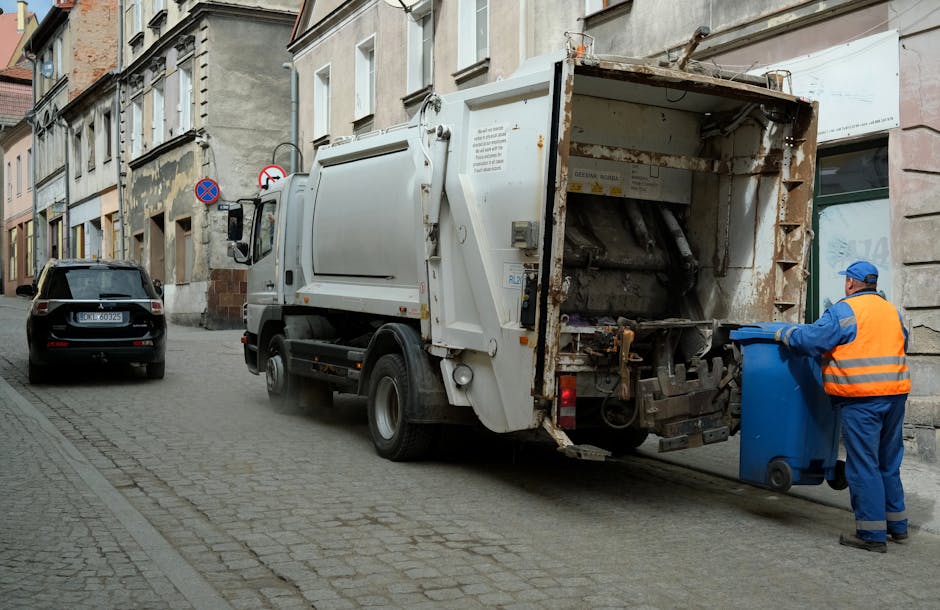 A waste collection vehicle parked on a narrow urban street during daytime, with its rear hatch open and visible mechanical components used for compacting rubbish. To the right, a worker dressed in blue overalls and a high-visibility orange vest, with a blue cap, is pushing a blue wheelie bin toward the vehicle. The street is paved with cobblestones, lined with multi-storey buildings featuring a mix of stone and brick facades, with some windows and exterior signage visible. A parked black car is seen further along the street on the left side, and road signs indicating parking restrictions are mounted on the building wall. The scene, illuminated by natural daylight, captures a typical urban waste collection operation which may involve private or independent rubbish removal services, such as those offered by Waste Removal Knightsbridge, providing on-site clearance of general waste materials in the vicinity of Sloane Street and Knightsbridge. The environment is clean, with no visible debris on the street surface, and the overall atmosphere suggests a routine, professional rubbish collection process. This detailed visual description supports understanding of private waste handling methods in a city setting. 