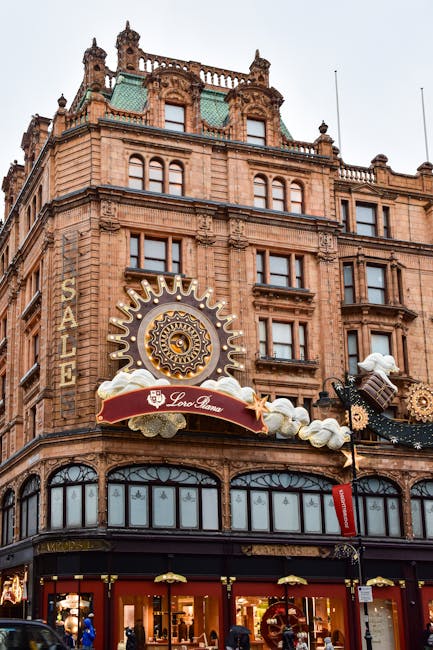 The image depicts the facade of a historic multi-storey building constructed from reddish-brown stone, featuring ornate architectural details such as decorative columns, cornices, and small turrets at the roofline. Large windows with arched and rectangular shapes are visible on each level, with some windows having decorative framing. The ground floor hosts retail storefronts with large glass display windows, showcasing interior merchandise. Prominently affixed to the upper portion of the building is a decorative, circular clock-like ornament with gold accents, surrounded by cloud motifs made of painted or sculpted material. Below the ornament, a red and gold sign bearing the name 'Loro Piana' extends outward, supported by decorative brackets. Adjacent to the sign, black street lamps and hanging decorative elements are visible, contributing to a festive or themed atmosphere. The scene is illuminated by natural daylight, revealing the intricate details of the building’s facade and the bustling retail environment at street level. This scene aligns with private waste handling activities, common in commercial areas where independent collection services such as Waste Removal Knightsbridge might operate to support ongoing maintenance and clearance of rubbish and waste from retail premises.